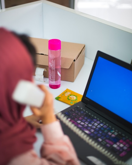 A person with a headscarf is on the phone at a desk. The workspace includes a laptop with a blue screen, a pink bottle, a cardboard box, and a packet of snacks. The person is facing away, focusing on the phone call.