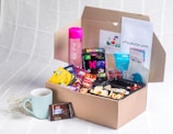 A cardboard box filled with various snacks, teas, and a pink bottle. The box is placed on a textured white surface. There is also a light blue mug beside the box, a dark chocolate bar, and some documents with writing.