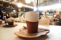 A warm cafe setting features a ceramic cup on a handcrafted wooden coaster. The background reveals an eclectic mix of furniture and decor, with soft lighting casting a cozy ambiance. A spoon rests beside the cup, and another beverage is visible in the background, hinting at a social setting.