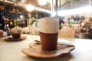 A warm cafe setting features a ceramic cup on a handcrafted wooden coaster. The background reveals an eclectic mix of furniture and decor, with soft lighting casting a cozy ambiance. A spoon rests beside the cup, and another beverage is visible in the background, hinting at a social setting.