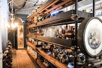 A narrow, industrial-looking room filled with shelves displaying an assortment of vintage metalware, such as kettles and pots. On the left side, a chandelier illuminates the space, casting warm light on the items. The walls and ceiling feature exposed metal piping, contributing to the rustic feel. At the far end, there's a doorway leading to another room.