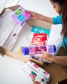A person is unpacking a cardboard box containing various items including a pack of feminine hygiene pads, a bottle of lotion or body spray, a bag of snacks, and a floral patterned pouch. The person is wearing a blue top and has their fingernails painted black.