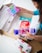 A person is unpacking a cardboard box containing various items including a pack of feminine hygiene pads, a bottle of lotion or body spray, a bag of snacks, and a floral patterned pouch. The person is wearing a blue top and has their fingernails painted black.