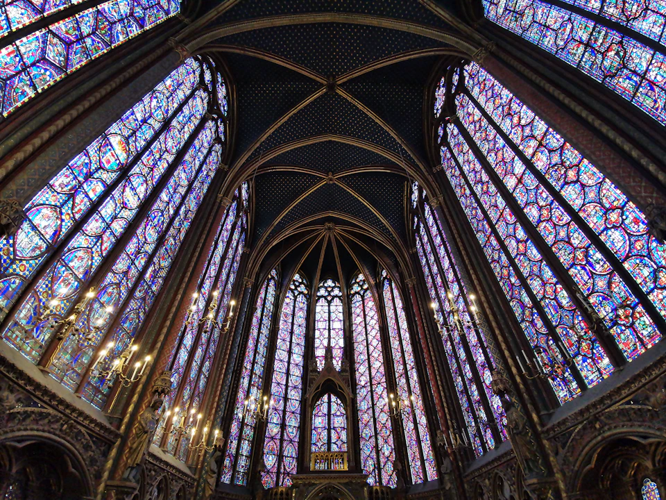 The image depicts a grand, Gothic-style church with tall, pointed arches and stained glass windows that fill the interior with vibrant colors. The high ceiling is supported by stone columns, adding to the architectural beauty of the space. The perspective of the photo is from below, looking up towards the towering structure, emphasizing its height and majesty.