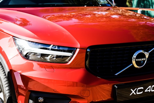 A close-up of the front section of a red car, showcasing a modern headlight design and a prominent logo in the center of the glossy black grille. The curvature of the bodywork reflects the surrounding environment, emphasizing the sleek and stylish appearance.