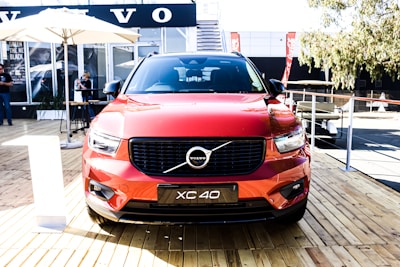 A red Volvo XC40 is parked on a wooden platform in an outdoor setting. The car's design is modern and sleek, with prominent headlights and grille. In the background, there are banners and a building with glass windows. Umbrellas and people sitting or standing can be seen, indicating a casual, outdoor event.