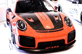 A sleek, high-performance sports car with a vibrant orange and black exterior is prominently displayed indoors. The car features aerodynamic lines and a distinctive front design with prominent headlights. The model identifier 'GT2RS' is visible on the front bumper.