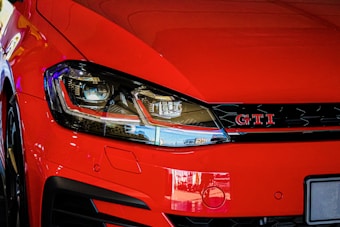 A close-up view of the front end of a red car featuring a prominent headlight and a GTI badge on the grille. The paint reflects the surrounding environment, creating a glossy effect.
