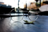 A pair of delicate round eyeglasses sits on a dark, reflective surface. The background is softly blurred, suggesting an indoor setting with light filtering through windows. A single green leaf is positioned beneath the glasses, adding a touch of nature to the composition.