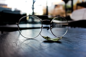A pair of delicate round eyeglasses sits on a dark, reflective surface. The background is softly blurred, suggesting an indoor setting with light filtering through windows. A single green leaf is positioned beneath the glasses, adding a touch of nature to the composition.