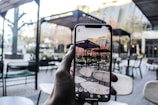 A hand is holding a smartphone displaying a live view of an outdoor patio area with chairs and tables. The background shows the same setting seen on the smartphone screen, featuring pergolas, chairs, and tables. The colors are muted with a mix of browns, beiges, and greens.