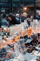 A bustling market scene with a variety of dried fruits and nuts in plastic bags displayed on a table. A woman is focused on her work behind the stall. In the background, people are gathered near the wire-mesh fencing with warm lighting, suggesting a busy atmosphere.