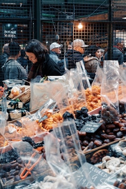 A bustling market scene with a variety of dried fruits and nuts in plastic bags displayed on a table. A woman is focused on her work behind the stall. In the background, people are gathered near the wire-mesh fencing with warm lighting, suggesting a busy atmosphere.