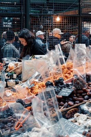 A bustling market scene with a variety of dried fruits and nuts in plastic bags displayed on a table. A woman is focused on her work behind the stall. In the background, people are gathered near the wire-mesh fencing with warm lighting, suggesting a busy atmosphere.