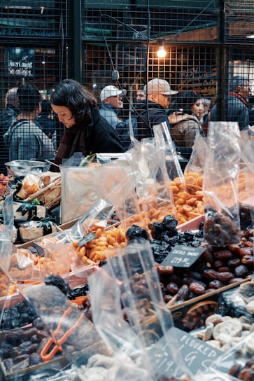 A bustling market scene with a variety of dried fruits and nuts in plastic bags displayed on a table. A woman is focused on her work behind the stall. In the background, people are gathered near the wire-mesh fencing with warm lighting, suggesting a busy atmosphere.