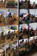 A collection of folded and rolled fabrics on display shelves, showcasing a variety of colors and patterns. The fabrics include checks, stripes, and solid colors, neatly organized on several wooden shelves with metal supports.