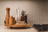 A kitchen countertop features a wooden cutting board holding a variety of kitchen items, including pepper mills, olive oil dispensers, and a small wooden container. The scene is set against a background of tiled wall with a neutral color palette.