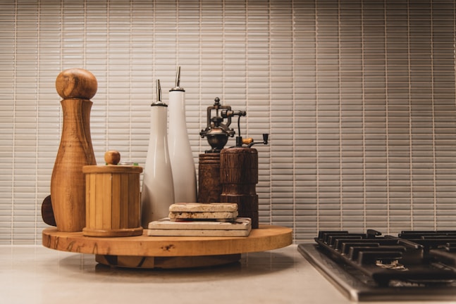A kitchen countertop features a wooden cutting board holding a variety of kitchen items, including pepper mills, olive oil dispensers, and a small wooden container. The scene is set against a background of tiled wall with a neutral color palette.