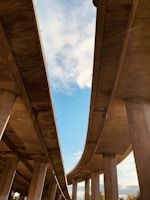 Two large concrete highways extend upwards towards the sky, creating a sharp contrast with the light blue sky and scattered white clouds. The perspective from underneath highlights the expansive structure of the highways, supported by thick pillars. The open sky visible between the highways adds depth and openness to the scene.