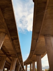 Two large concrete highways extend upwards towards the sky, creating a sharp contrast with the light blue sky and scattered white clouds. The perspective from underneath highlights the expansive structure of the highways, supported by thick pillars. The open sky visible between the highways adds depth and openness to the scene.