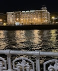 A beautifully lit hotel building at night, named Hotel Maria Cristina, stands prominently against the backdrop of a dark sky. The building's facade is illuminated by warm yellow lights that reflect off the rippling water in front. An ornate white metal railing is visible in the foreground, adding to the scene's elegance.