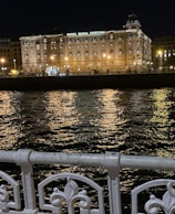 A beautifully lit hotel building at night, named Hotel Maria Cristina, stands prominently against the backdrop of a dark sky. The building's facade is illuminated by warm yellow lights that reflect off the rippling water in front. An ornate white metal railing is visible in the foreground, adding to the scene's elegance.