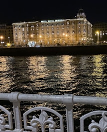 A beautifully lit hotel building at night, named Hotel Maria Cristina, stands prominently against the backdrop of a dark sky. The building's facade is illuminated by warm yellow lights that reflect off the rippling water in front. An ornate white metal railing is visible in the foreground, adding to the scene's elegance.