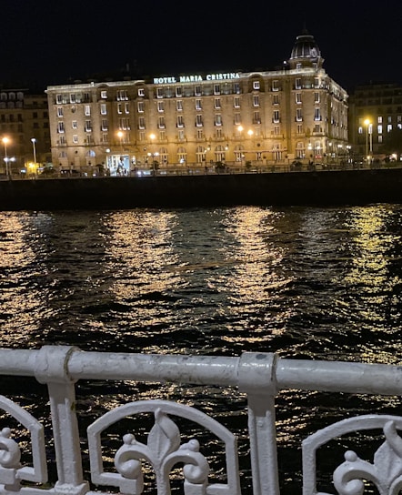 A beautifully lit hotel building at night, named Hotel Maria Cristina, stands prominently against the backdrop of a dark sky. The building's facade is illuminated by warm yellow lights that reflect off the rippling water in front. An ornate white metal railing is visible in the foreground, adding to the scene's elegance.