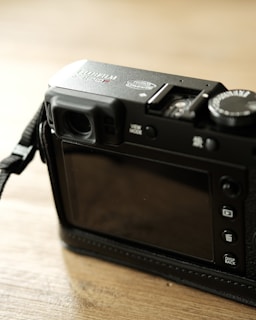 A digital camera is positioned on a wooden surface. The focus is on the back of the camera, displaying a screen and several buttons. The camera is predominantly black and features a textured body.