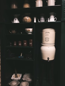 A dark wooden cabinet filled with vintage kitchenware and ceramic jars. The upper shelves contain various pitchers, bowls, and jugs, some with floral designs. The lower section holds labeled canisters with French inscriptions, a mortar and pestle, and a large ceramic jar with a spigot.