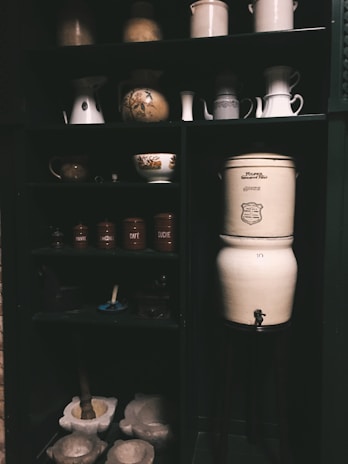 A dark wooden cabinet filled with vintage kitchenware and ceramic jars. The upper shelves contain various pitchers, bowls, and jugs, some with floral designs. The lower section holds labeled canisters with French inscriptions, a mortar and pestle, and a large ceramic jar with a spigot.