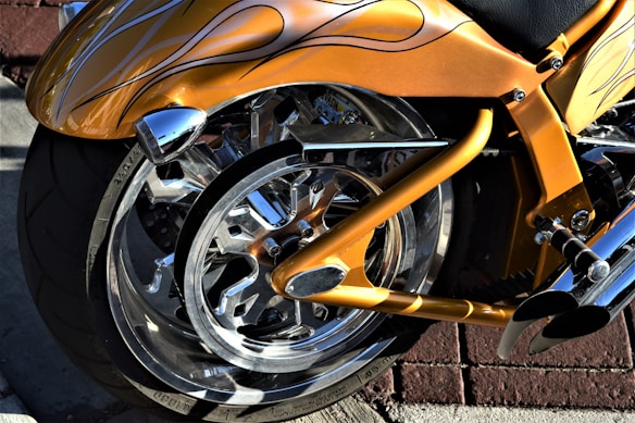 A close-up view of a custom motorcycle with a striking metallic orange paint job featuring flame designs. The image focuses on the rear wheel, showcasing shiny chrome elements and the intricate design of the wheel and frame. The background consists of a red brick wall and pavement.