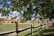 A rural scene with a farm setting highlighted by a group of people gathered around a large stack of hay bales. The background includes a white domed structure, and the scene is bordered by lush green trees and a wooden fence. The path is lined with wooden stakes, and there are individuals walking and interacting in the area.