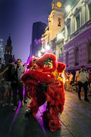 A vibrant red lion costume is displayed prominently in a nighttime street performance, surrounded by a crowd of onlookers. The background showcases an illuminated historical building with a clock tower, adding an urban and festive atmosphere.