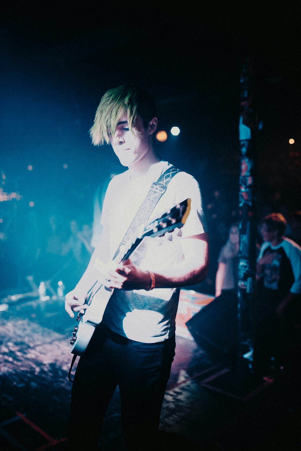 man wearing white crew-neck t-shirt standing while playing guitar surrounded with people watching