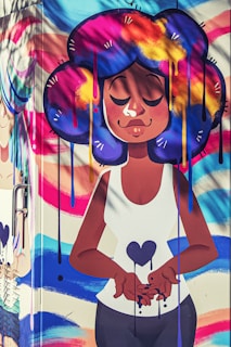 A vibrant street art mural featuring a cartoon-style character with large, colorful hair in shades of blue, purple, yellow, and pink. The character is depicted with closed eyes, expressing serenity, and holding a heart symbol. The background includes swirling, colorful patterns.