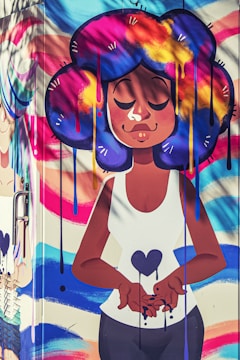 A vibrant street art mural featuring a cartoon-style character with large, colorful hair in shades of blue, purple, yellow, and pink. The character is depicted with closed eyes, expressing serenity, and holding a heart symbol. The background includes swirling, colorful patterns.