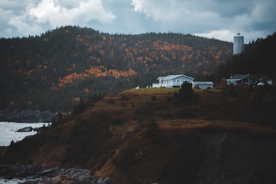 A serene landscape featuring a house on a grassy hill overlooking a body of water. Surrounding the house are autumn-colored trees with hues of orange and yellow. In the background, a mountainous region is visible under a cloudy sky, and a tall structure stands to the right of the house.