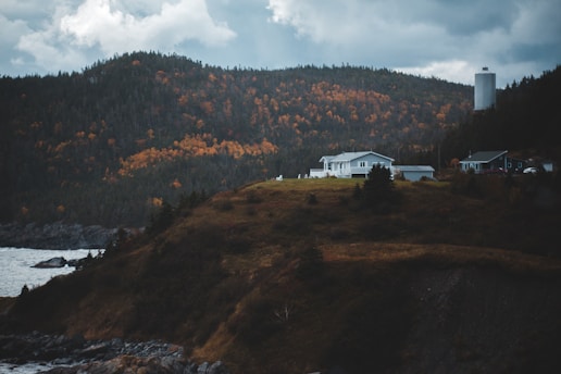 A serene landscape featuring a house on a grassy hill overlooking a body of water. Surrounding the house are autumn-colored trees with hues of orange and yellow. In the background, a mountainous region is visible under a cloudy sky, and a tall structure stands to the right of the house.