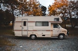 A vintage motorhome parked on a gravel path surrounded by autumn trees. The vehicle has a cream color with brown stripes and the name 'Centurion' emblazoned on its side. The setting suggests a quiet, rural area with leafy trees showcasing orange and yellow foliage.