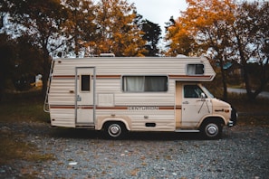 A vintage motorhome parked on a gravel path surrounded by autumn trees. The vehicle has a cream color with brown stripes and the name 'Centurion' emblazoned on its side. The setting suggests a quiet, rural area with leafy trees showcasing orange and yellow foliage.