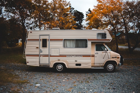 A vintage motorhome parked on a gravel path surrounded by autumn trees. The vehicle has a cream color with brown stripes and the name 'Centurion' emblazoned on its side. The setting suggests a quiet, rural area with leafy trees showcasing orange and yellow foliage.