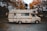 A vintage motorhome parked on a gravel path surrounded by autumn trees. The vehicle has a cream color with brown stripes and the name 'Centurion' emblazoned on its side. The setting suggests a quiet, rural area with leafy trees showcasing orange and yellow foliage.