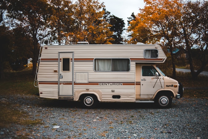 A vintage motorhome parked on a gravel path surrounded by autumn trees. The vehicle has a cream color with brown stripes and the name 'Centurion' emblazoned on its side. The setting suggests a quiet, rural area with leafy trees showcasing orange and yellow foliage.