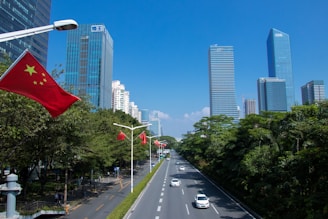 A bustling urban scene with skyscrapers lining both sides of a wide road. Several Chinese flags are mounted on lamp posts along the road. The sky is clear and blue, and there is greenery on one side of the street.