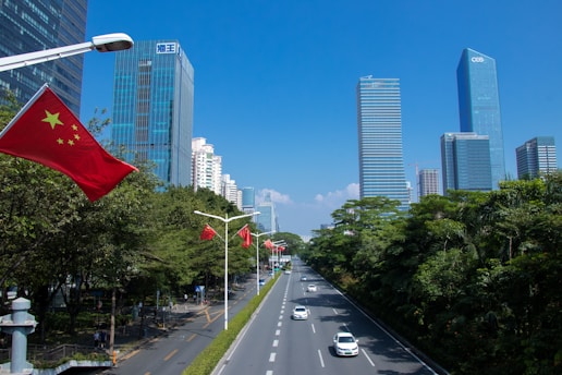 A bustling urban scene with skyscrapers lining both sides of a wide road. Several Chinese flags are mounted on lamp posts along the road. The sky is clear and blue, and there is greenery on one side of the street.