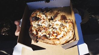 A partially eaten pizza with a crispy, charred crust is placed in a cardboard takeout box. The pizza is topped with melted cheese and mushrooms. The box is being held by a person whose hands are visible.