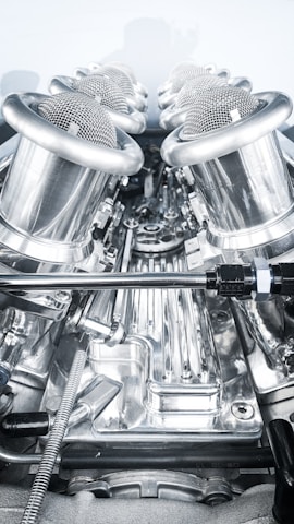 A close-up view of a complex, polished metallic engine component, featuring a series of cylindrical, mesh-covered air intake manifolds. The metal surfaces are reflective and intricately designed, indicating high precision engineering.