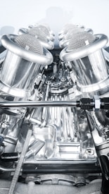 A close-up view of a complex, polished metallic engine component, featuring a series of cylindrical, mesh-covered air intake manifolds. The metal surfaces are reflective and intricately designed, indicating high precision engineering.