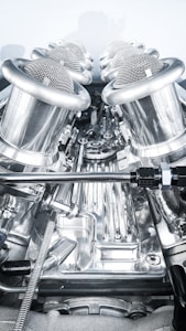 A close-up view of a complex, polished metallic engine component, featuring a series of cylindrical, mesh-covered air intake manifolds. The metal surfaces are reflective and intricately designed, indicating high precision engineering.